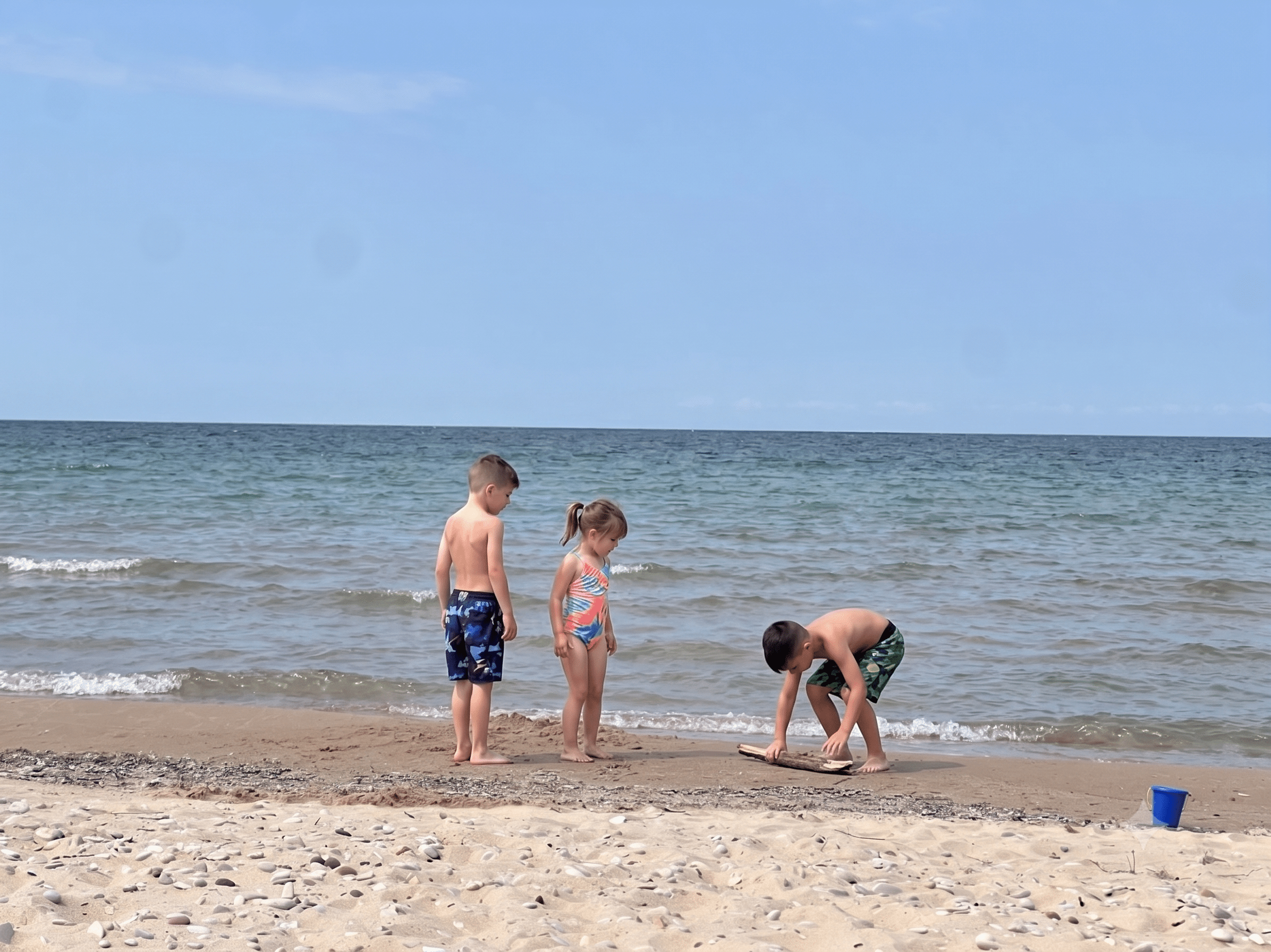 Children playing at a sandy beach along Michigan’s Tunnel of Trees route, digging at the shoreline beside calm lake water on a sunny day.
